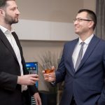 Two business professionals socializing indoors, wearing formal attire, engaging in a friendly conversation.